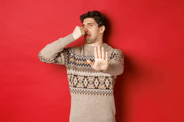悪い匂いで何かを拒否し鼻を閉じて手を上げる冬のセーターのうんざりハンサムな男のイメージは、赤の背景の上に立って — ストック写真