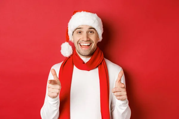 Konzept von Weihnachten, Winterurlaub und Feiern. Frecher Mann mit Weihnachtsmütze und Schal, lächelnd und mit den Fingern in die Kamera zeigend, frohes neues Jahr wünschend, vor rotem Hintergrund stehend — Stockfoto
