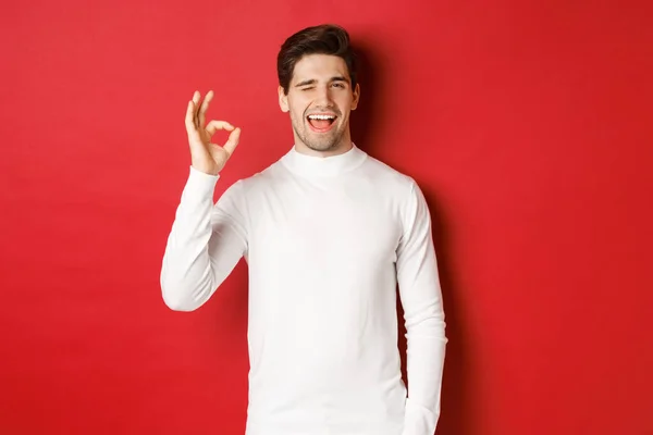 Concept de vacances d'hiver, Noël et style de vie. Un bel homme en pull blanc qui vous fait un clin d'œil, qui montre un signe et un sourire acceptables, qui garantit la qualité, qui recommande quelque chose — Photo