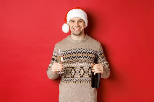 Begrip wintervakantie, nieuwjaar en viering. Portret van een knappe man in kerstmuts en trui, champagne vasthoudend en toast makend op kerstfeest, staande over rode achtergrond — Stockfoto