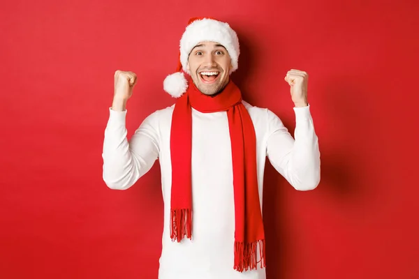 Портрет счастливого и взволнованного человека в шляпе и шарфе Санты, радующегося и выигрывающего что-то, празднующего новый год, стоящего на красном фоне — стоковое фото