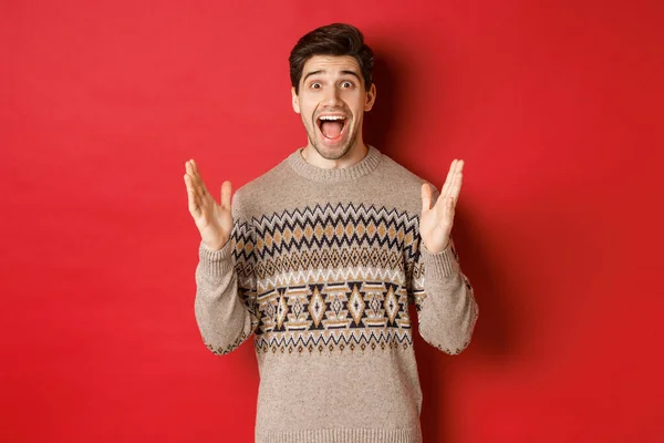 Portrait of surprised and happy handsome man, wearing christmas sweater, looking amazed, celebrating new year, standing over red background — Stock Photo, Image