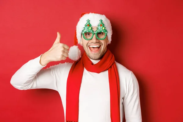 Koncepcja Bożego Narodzenia, ferie zimowe i uroczystości. Wesoły facet w czapce Mikołaja i okularach imprezowych, z kciukiem w górze i uśmiechem zadowolony, stojący na czerwonym tle — Zdjęcie stockowe