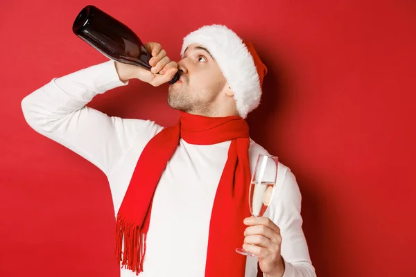 Concept of winter holidays, christmas and lifestyle. Man getting drunk on new year party, drinking champagne from bottle, wearing santa hat, standing over red background — Stock Photo, Image