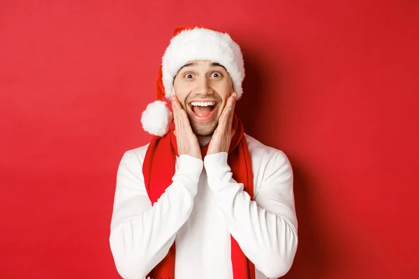 Conceito de Natal, férias de inverno e celebração. Close-up de homem surpreso e feliz em santa chapéu e cachecol, olhando para algo incrível, de pé sobre fundo vermelho — Fotografia de Stock