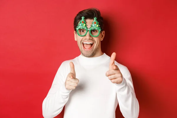 Joyful caucasian guy in party glasses and white sweater, smiling and pointing fingers at camera, wishing merry christmas and happy new year, standing over red background — Stock Photo, Image