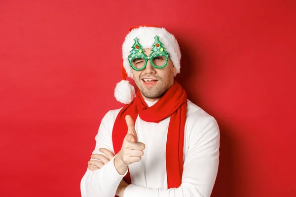 Konzept von Weihnachten, Winterurlaub und Feiern. Großaufnahme eines frechen jungen Mannes mit Weihnachtsmannmütze und Partybrille, der lächelnd mit dem Finger in die Kamera zeigt und über rotem Hintergrund steht — Stockfoto