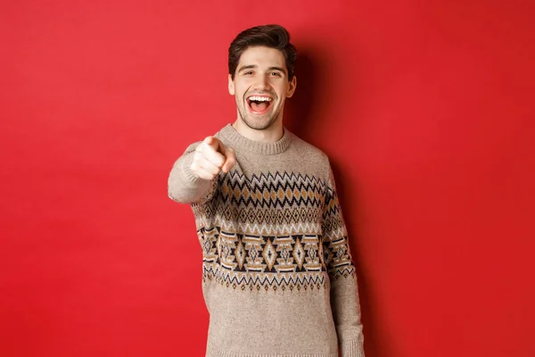 Concept of christmas celebration, winter and lifestyle. Happy attractive man in xmas sweater, wishing happy holidays and pointing at camera, standing over red background — Stock Photo, Image