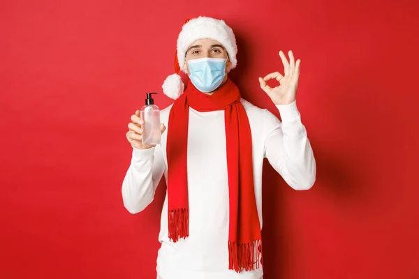 Begreppet covid-19, jul och helgdagar under pandemin. Attraktiv man i Santa hatt och medicinsk mask, visar okej tecken samtidigt rekommendera handdesinfektionsmedel, står över röd bakgrund — Stockfoto