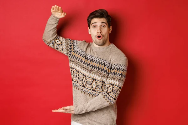 Afbeelding van een aantrekkelijke man in kerst trui, vormgeving van grote nieuwjaar cadeau, toont iets groots en verbazingwekkend, staande over rode achtergrond — Stockfoto