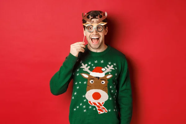 Красива здивована людина в різдвяному светрі, тримає партійну маску і посміхається, насолоджуючись святкуванням нового року, стоїть на червоному тлі — стокове фото