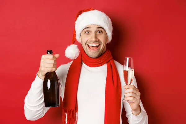 Begreppet vinter semester, jul och livsstil. Närbild av glada stilig man, håller champagne flaska och glas, firar nytt år, står över röd bakgrund — Stockfoto
