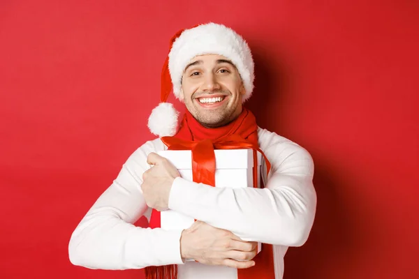 Concetto di vacanze invernali, Natale e stile di vita. Immagine di bel ragazzo in cappello di Babbo Natale e sciarpa, abbracciando il suo nuovo anno presente e sorridente lusingato, in piedi su sfondo rosso — Foto Stock