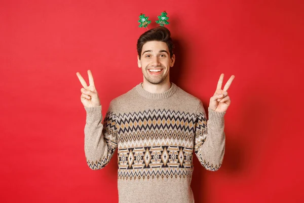 Koncept zimních prázdnin, vánoc a oslav. Obrázek pohledného a hloupého chlapa oblečeného na novoroční párty, ukazující mírové značky a usmívající se, stojící na červeném pozadí — Stock fotografie