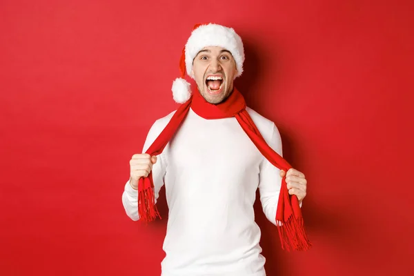 Afbeelding van boze volwassen man haat kerst, het dragen van sjaal en santa hoed, schreeuwen verontwaardigd, staande boos over rode achtergrond — Stockfoto