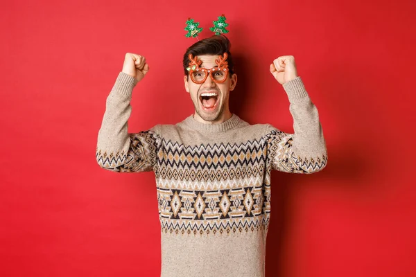 Портрет взволнованного и счастливого человека в бокалах для вечеринок, зимний свитер, крик радости и поднятие рук, выигрыш приза на Рождество, достижение цели нового года, стоя на красном фоне — стоковое фото