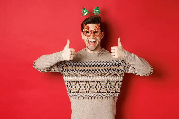 Noel süveteri ve parti gözlükleri içinde mutlu ve mutlu bir adamın portresi. Başparmaklarını göstererek mutlu yıllar diliyor. Kırmızı arka planda dikiliyor. — Stok fotoğraf