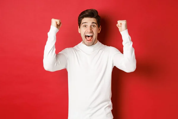Retrato de homem feliz animado em camisola branca, levantando as mãos para cima e triunfando, celebrando o ano novo, de pé feliz contra o fundo vermelho — Fotografia de Stock