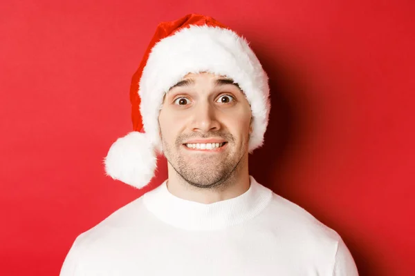 Gros plan d'un beau mec au chapeau de Père Noël, mordant la lèvre et regardant avec tentation quelque chose qu'il veut, debout sur fond rouge — Photo