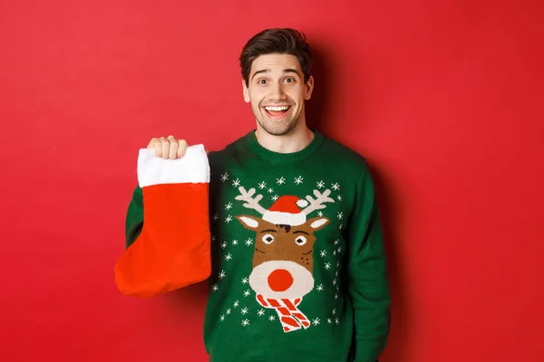 Imagen de chico feliz guapo en suéter, sosteniendo medias de Navidad y sonriendo divertido, de pie sobre fondo rojo —  Fotos de Stock