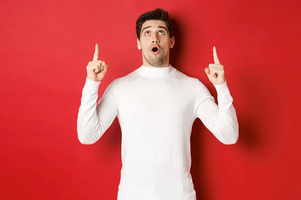 Begreppet vintersemester. Porträtt av stilig överraskad man i vit tröja, reagerar på nyår promo erbjudande, tittar och pekar finger upp, stående imponerad mot röd bakgrund — Stockfoto