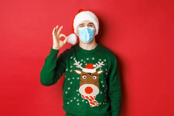 Концепція нового року, коїд-19 та соціальне дистанціювання. Веселий чоловік в медичній масці і капелюсі Санта, показуючи добре знак, рекомендую щось хороше, стоячи на червоному тлі — стокове фото