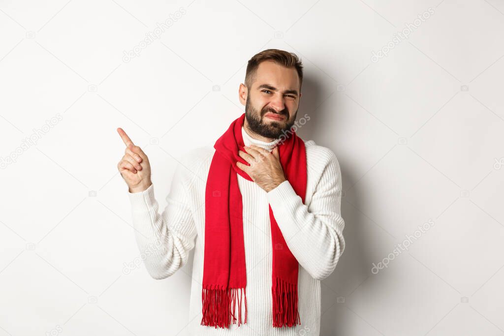 Christmas shopping and winter holidays concept. Sick bearded man with sore throat pointing finger at upper left corner, need medication, standing over white background