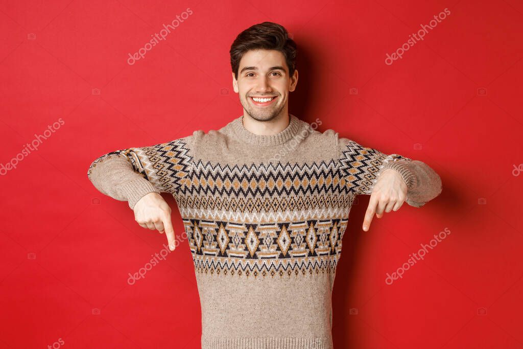Concept of christmas celebration, winter holidays and lifestyle. Attractive happy man in xmas sweater showing promotion, pointing fingers down at logo, standing over red background.