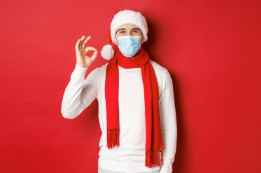 Covid-19, noel ve salgın sırasında tatil kavramı. Tıbbi maske ve Noel Baba şapkası takan mutlu ve memnun bir adamın portresi onay işareti gösteriyor, bir şey öneriyor.