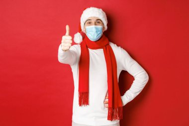 Covid-19, noel ve salgın sırasında tatil kavramı. Tıbbi maske ve Noel Baba şapkası takan neşeli yakışıklı adam, baş parmağını kaldırıyor, yeni yılı ve sosyal mesafeyi kutluyor.