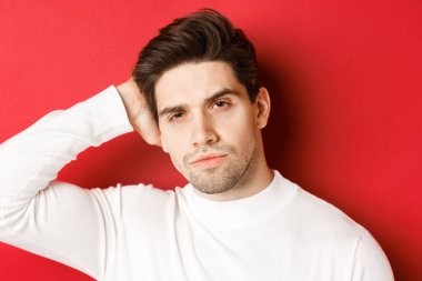 Yakışıklı esmer adamın saç kesimine dokunduğu, kameraya küstah baktığı, kırmızı arka planda beyaz kazak giydiği yakın plan.