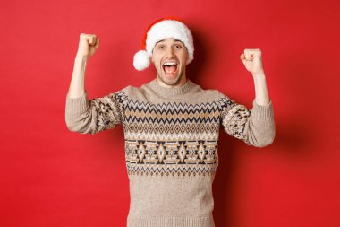 Küfürbaz ve Noel Baba şapkalı neşeli yakışıklı bir adam imajı, yeni yılı kutluyor, bir şey kazanıyor ya da kazanıyor, ellerini kaldırıp sevinç çığlıkları atıyor, kırmızı arka planda duruyor.