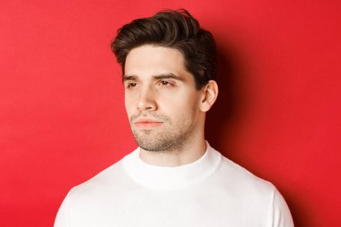 Beyaz kazaklı, ciddi ve düşünceli bir adamın yakın çekimi, sola bakıyor, kırmızı arka planda duruyor.