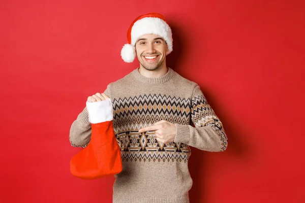 Kış tatili, yeni yıl ve kutlama kavramı. Yakışıklı gülümseyen adam çocuklar için hediyeler hazırladı. Yılbaşı çorap çantasına işaret etti. Kırmızı arka planda duruyordu. — Stok fotoğraf