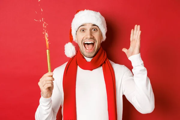 Konzept von Weihnachten, Winterurlaub und Feiern. Schöner Mann feiert Neujahr und hat Spaß, hält Wunderkerze in der Hand und lächelt, trägt Weihnachtsmütze, steht über rotem Hintergrund — Stockfoto