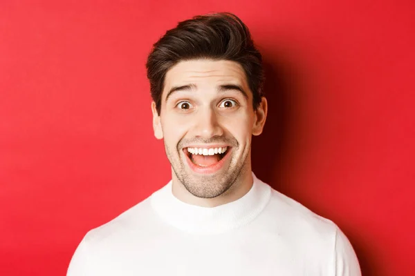 Nære på, overrasket, lykkelig fyr i hvit genser, ser underholdende ut, hører interessante nyheter, står over rød bakgrunn – stockfoto