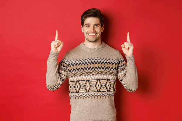 Portrait of attractive guy in christmas sweater pointing fingers up, smiling and showing winter holidays advertisement, celebrating new year over red background — Stock Photo, Image
