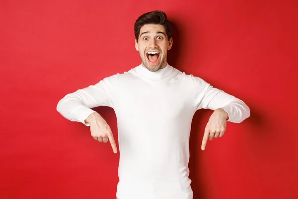 Εικόνα του έκπληκτος όμορφος άντρας σε λευκό πουλόβερ, αντιδρώντας ενθουσιασμένος με τη διαφήμιση των Χριστουγέννων, δείχνοντας τα δάχτυλα προς τα κάτω, δείχνει το λογότυπο, στέκεται πάνω από το κόκκινο φόντο — Φωτογραφία Αρχείου