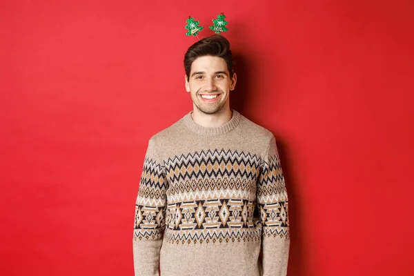 Concepto de vacaciones de invierno, Navidad y celebración. Chico barbudo guapo en suéter deseando feliz año nuevo, sonriendo a la cámara, de pie sobre el fondo rojo —  Fotos de Stock