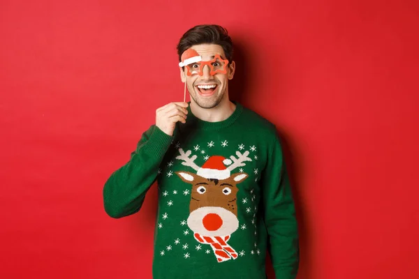 Портрет красивого усміхненого чоловіка в різдвяному светрі і вечірніх окулярах, виглядає здивованим і щасливим, святкуючи новий рік і розважаючись, стоїть на червоному тлі — стокове фото