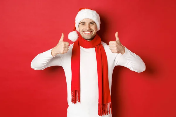 Porträtt av stilig leende man i Santa halsduk och hatt, visar tummen upp, firar jul, står över röd bakgrund — Stockfoto
