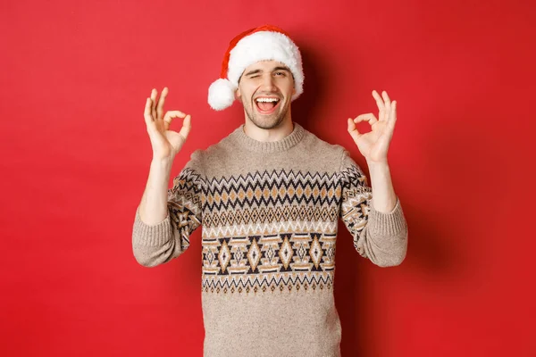 Εικόνα ενός χαρούμενου ελκυστικού άντρα με πουλόβερ και καπέλο santa, που εύχεται Καλά Χριστούγεννα, δείχνει εντάξει σημάδια και κλείνει το μάτι στην κάμερα, γιορτάζει το νέο έτος, στέκεται πάνω από το κόκκινο φόντο — Φωτογραφία Αρχείου