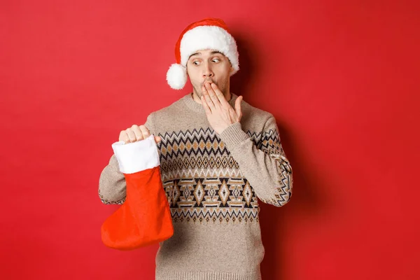 Konzept der Winterferien, Neujahr und Feier. Überraschter erwachsener Mann hält Weihnachtsstrumpf mit Geschenken drin, blickt erstaunt auf Tasche, steht über rotem Hintergrund in Weihnachtsmütze — Stockfoto