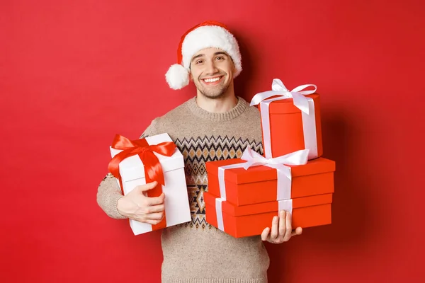 Έννοια των χειμερινών διακοπών, το νέο έτος και γιορτή. Πορτρέτο του αξιαγάπητου χαμογελαστού άνδρα ετοίμασε δώρα για τα Χριστούγεννα, κρατώντας δώρα και κοιτάζοντας εγκάρδια κάμερα, κόκκινο φόντο — Φωτογραφία Αρχείου