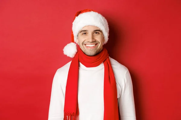 Conceito de férias de inverno, Natal e estilo de vida. Close-up de homem bonito com cerdas, vestindo chapéu de Natal com cachecol e sorrindo feliz, desejando feliz ano novo, fundo vermelho — Fotografia de Stock