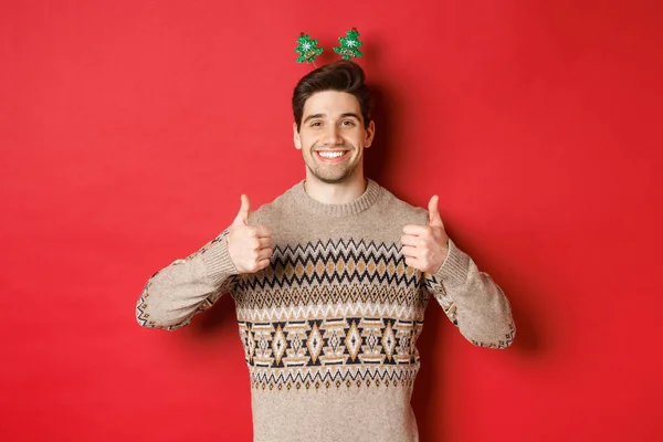 Conceito de férias de inverno, Natal e celebração. Cara barbuda alegre em suéter, mostrando polegares para cima na aprovação e sorrindo, desfrutando de festa de ano novo, fundo vermelho — Fotografia de Stock