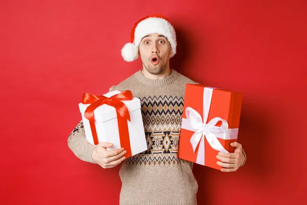Koncepcja ferii zimowych, Nowy Rok i świętowanie. Wizerunek zaskoczonego atrakcyjnego faceta w swetrze Mikołaja i Boże Narodzenie, otrzymującego prezenty, trzymającego prezenty i wyglądającego na zdumionego — Zdjęcie stockowe