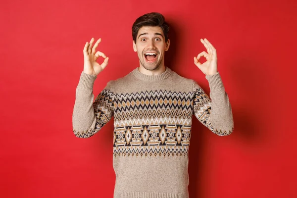 Begrip kerstfeest, wintervakantie en levensstijl. Portret van een knappe man in trui, die verbaasd kijkt en goede tekenen vertoont, iets goeds garandeert of aanbeveelt, rode achtergrond — Stockfoto