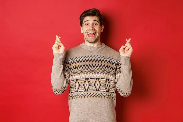 Imagem do jovem animado e esperançoso fazendo desejo, vestindo camisola de inverno, cruzando os dedos para boa sorte e sorrindo, antecipando véspera de Natal, de pé sobre fundo vermelho — Fotografia de Stock