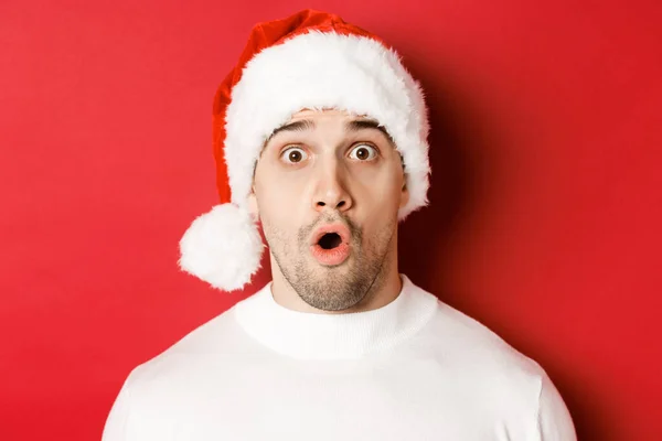 Närbild av attraktiv överraskad man i Santa hatt, säger wow och tittar på något intressant, står mot röd bakgrund — Stockfoto